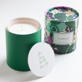 A green cylindrical scented candle with its white lid placed in front, and the matching floral-patterned tin canister standing behind it on a clean white background.