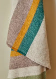Close up of pure new wool throw in a rich multistripe of olive, mustard, emerald green, and camel on a natural beige background.