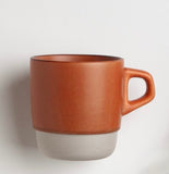 A ceramic mug by Kinto with a two-tone design, featuring a matte terracotta brown upper body and a light gray lower base, set against a plain light gray background.