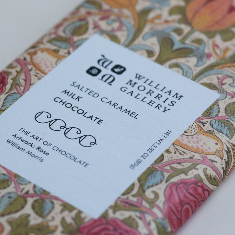 A close-up of a William Morris Gallery chocolate bar with a white paper label reading 'Salted Caramel Milk Chocolate' and 'William Morris Gallery.' The wrapper has a colorful floral and ornamental pattern in pink, blue, green, and beige tones.