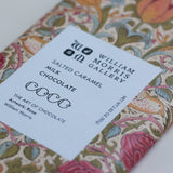 A close-up of a William Morris Gallery chocolate bar with a white paper label reading 'Salted Caramel Milk Chocolate' and 'William Morris Gallery.' The wrapper has a colorful floral and ornamental pattern in pink, blue, green, and beige tones.