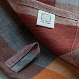 Close-up of a plaid linen dishtowel with a white fabric label reading 'LIVE LINEN' sewn onto it. The fabric features a muted plaid pattern in warm tones of red, orange and grey.