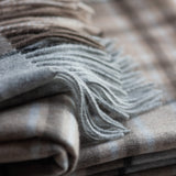 A close-up of a lambswool McKellar tartan  throw with fringed edges, shown in soft neutral tones of beige, taupe, and light gray.