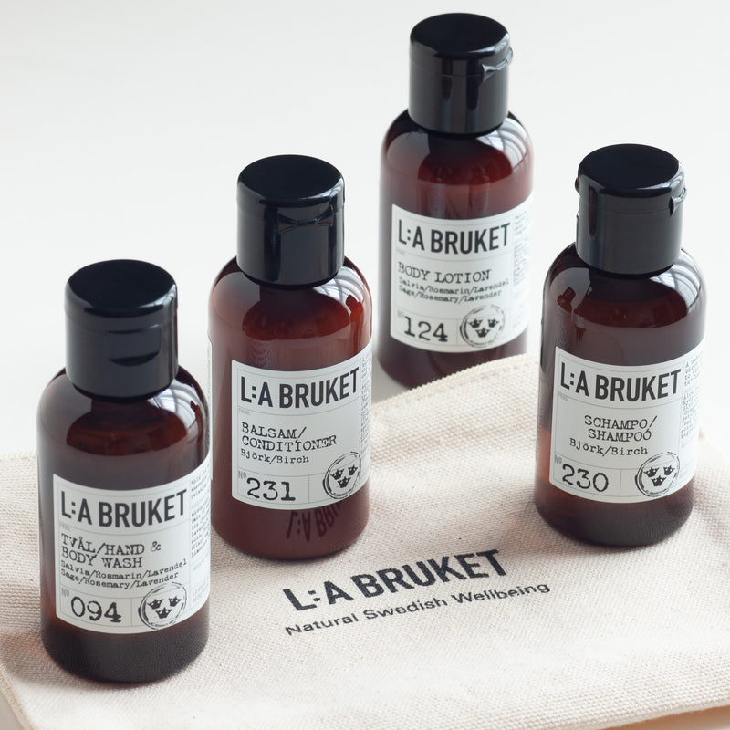 Four amber glass bottles of L'A Bruket natural Swedish wellbeing products arranged on a linen zip up bag The bottles include body wash, body lotion, shampoo, and conditioner.