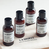 Four amber glass bottles of L'A Bruket natural Swedish wellbeing products arranged on a linen zip up bag The bottles include body wash, body lotion, shampoo, and conditioner.