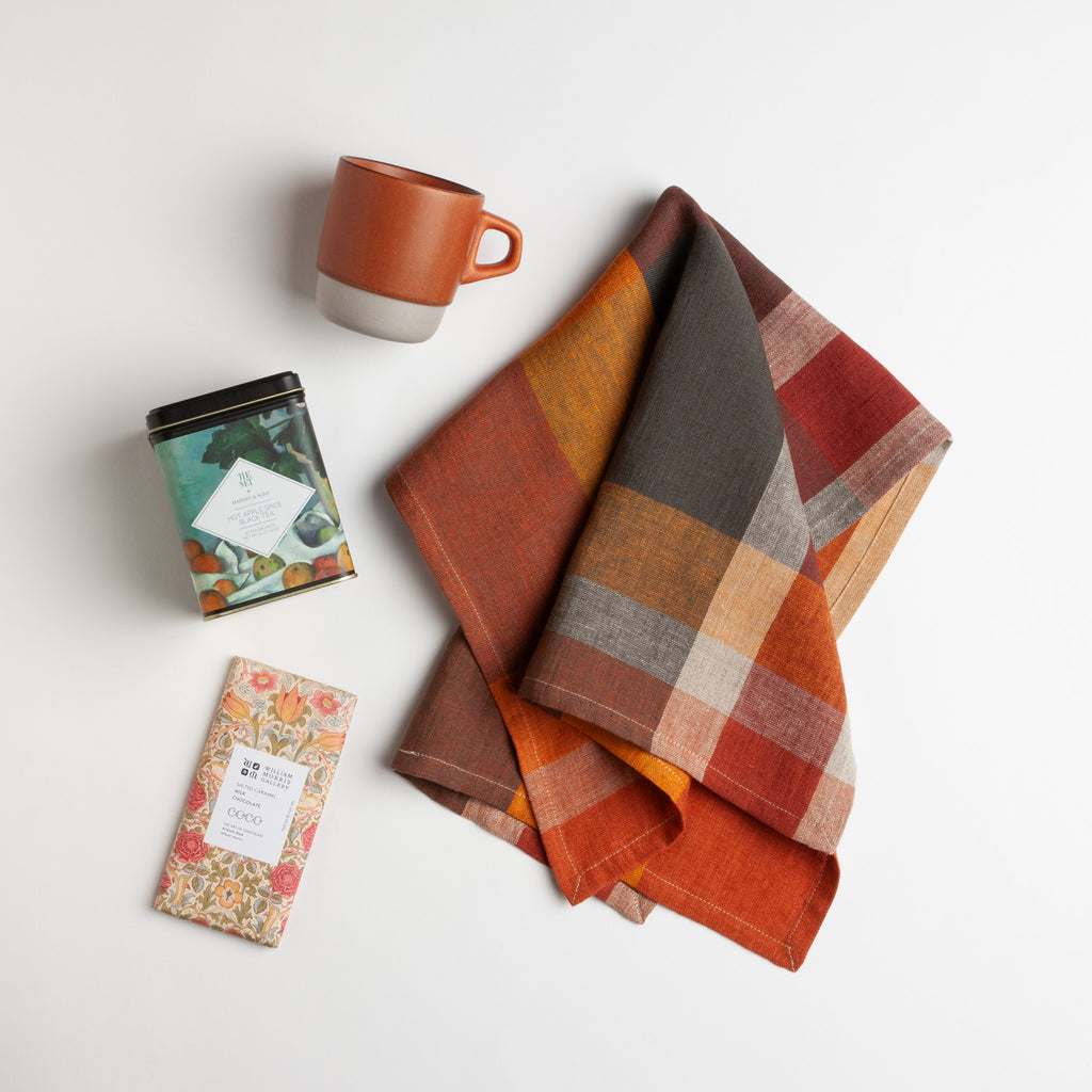 A folded plaid linen tea towel in warm autumn tones of orange, rust,and  brown,  styled on a white background with a terracotta mug, a apple spice tea, and a salted caramel milk chocolate bar.