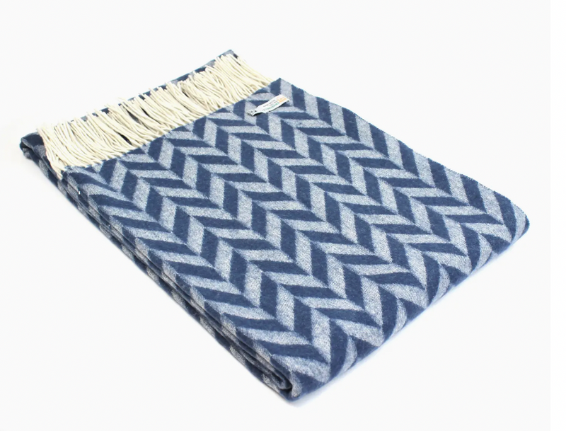 A folded lambswool throw blanket with a chunky blue and white herringbone pattern and cream fringe by Tweedmill. 
