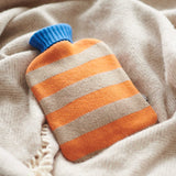Striped lambwool hot water bottle cover in orange and camel against the background of a beige herringbone blanket.