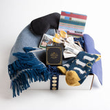 The star of this carton is a stunning teal and sandstone blanket with coordinating fringe. A collection of heritage teas, Smokey Bear playing cards, hydrating lip balm, a light-blocking silk eye mask, and lambswool socks adorned with countable sheep round out this distinctive get-well care package.
